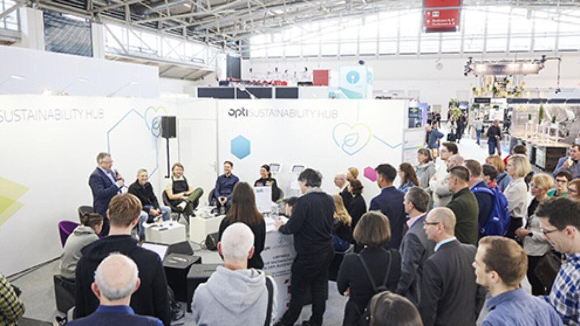 A group of people gathered at an indoor event, listening to a panel discussion at the Sustainability Hub section, with speakers seated on a stage and attendees standing and watching.