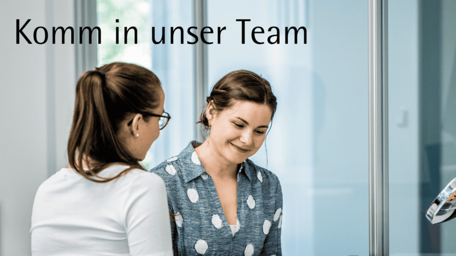 Two women are talking in an office with glass walls. One woman wears glasses, the other wears a blue polka dot shirt. The text Komm in unser Team appears at the top.