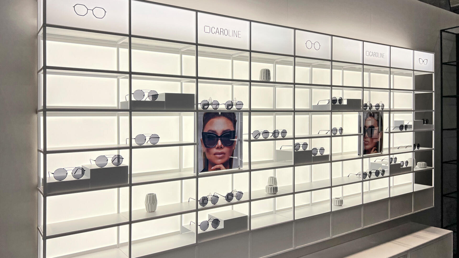 A modern, well-lit eyewear display wall with shelves holding various sunglasses and eyeglasses. There are two large photos of a woman wearing sunglasses, and the brand name CAROLINE is visible on illuminated signs.