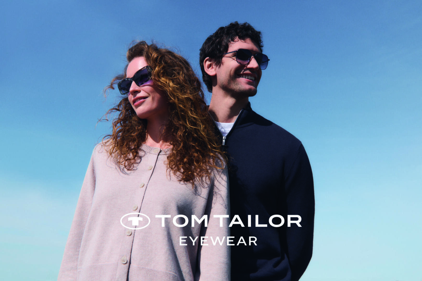 A woman and a man wearing sunglasses stand together outdoors under a clear blue sky. Both are smiling and looking into the distance. The text TOM TAILOR EYEWEAR appears at the bottom of the image.