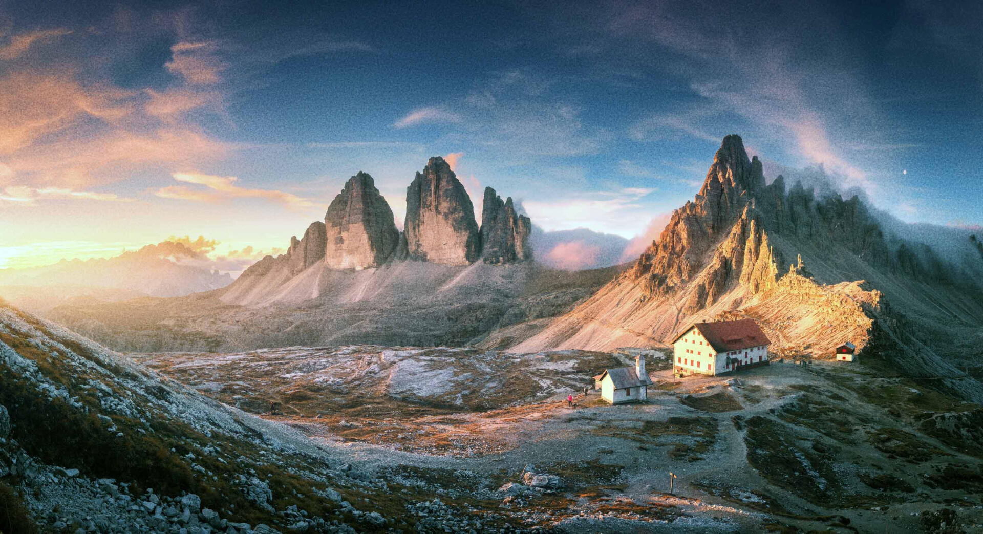 A panoramic view of jagged mountain peaks at sunrise, with sunlight illuminating a cluster of buildings in a rocky alpine landscape. The sky is partly cloudy, casting dramatic shadows on the scene.