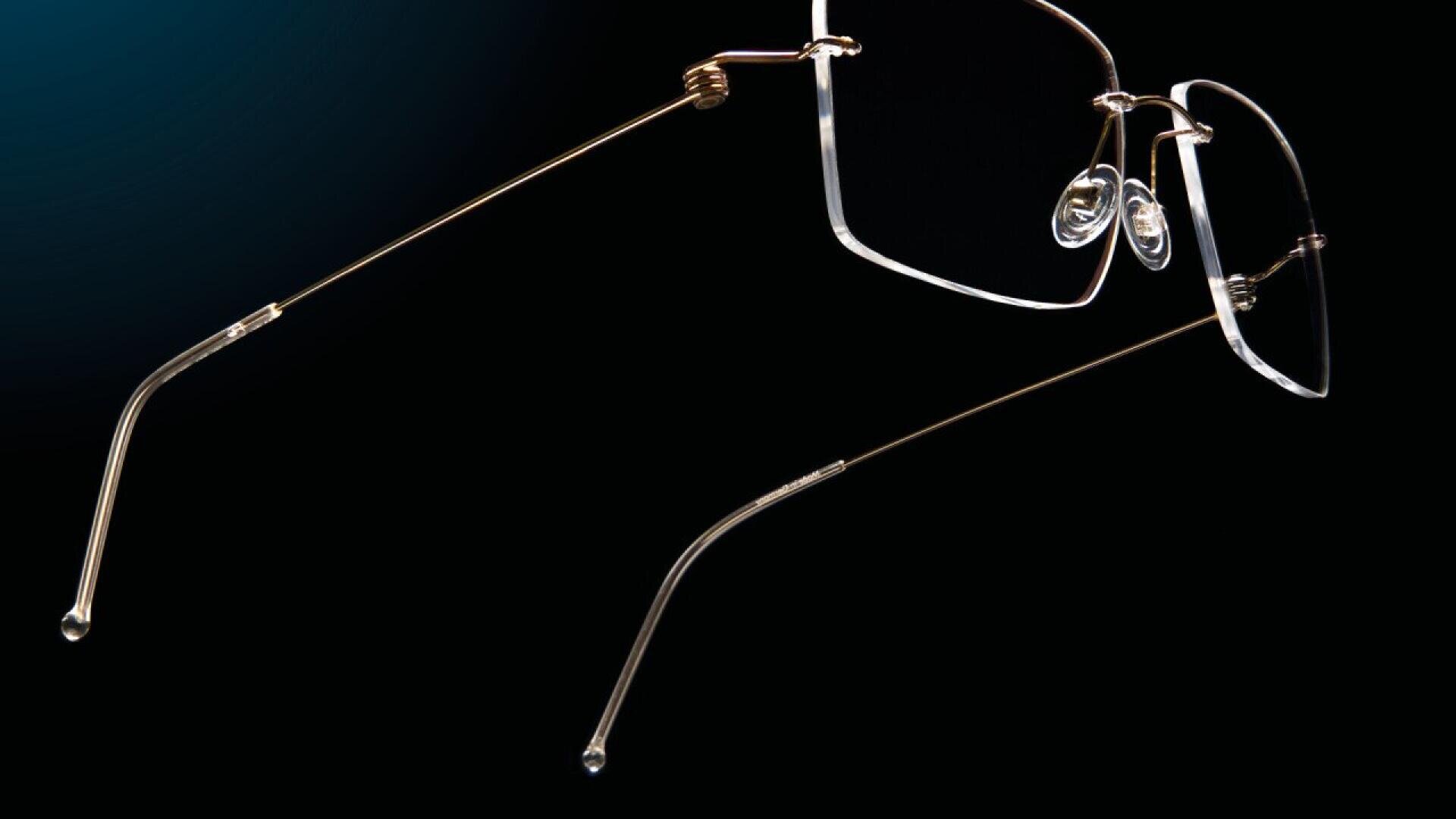A close-up of rimless eyeglasses with thin gold arms and clear nose pads, set against a black background.