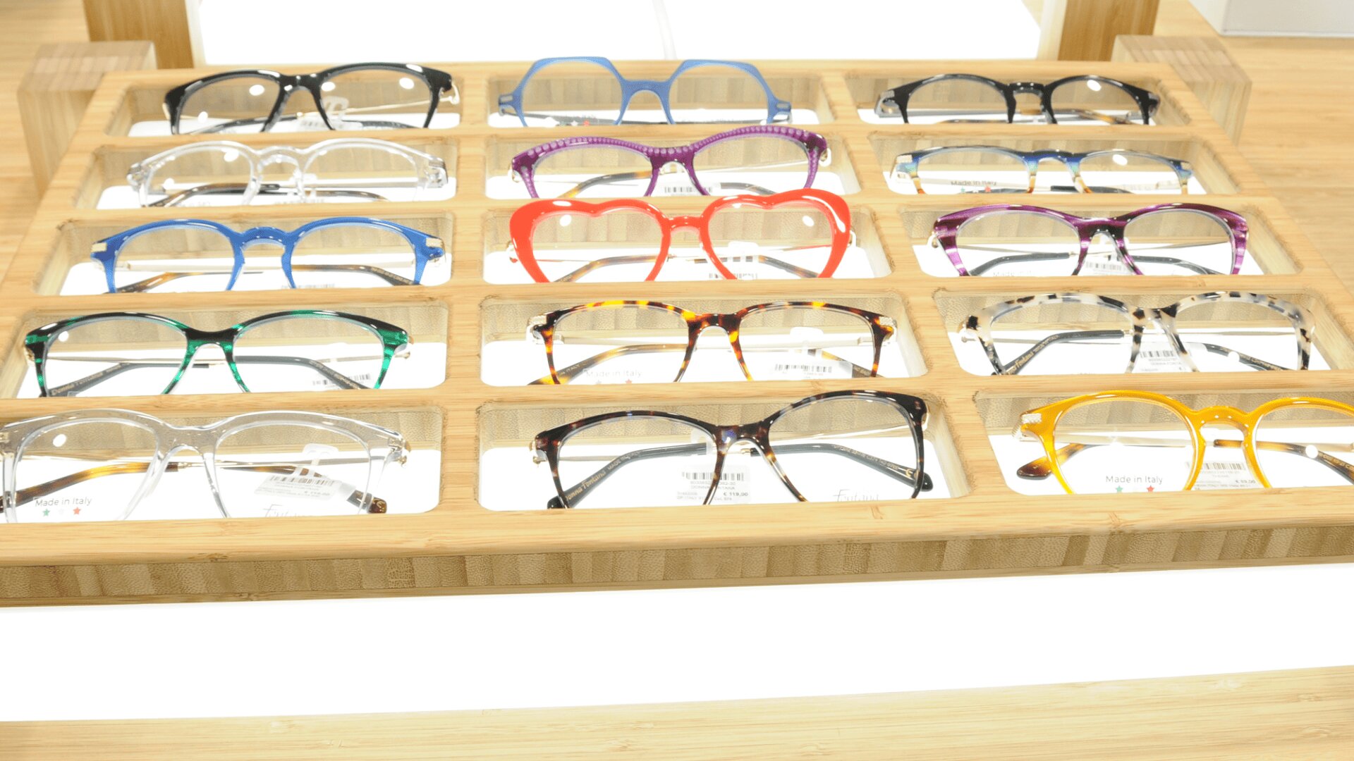 A wooden display tray with twelve pairs of eyeglasses in various colors and styles, including blue, red, purple, clear, tortoiseshell, black, green, and yellow, arranged neatly in three rows.