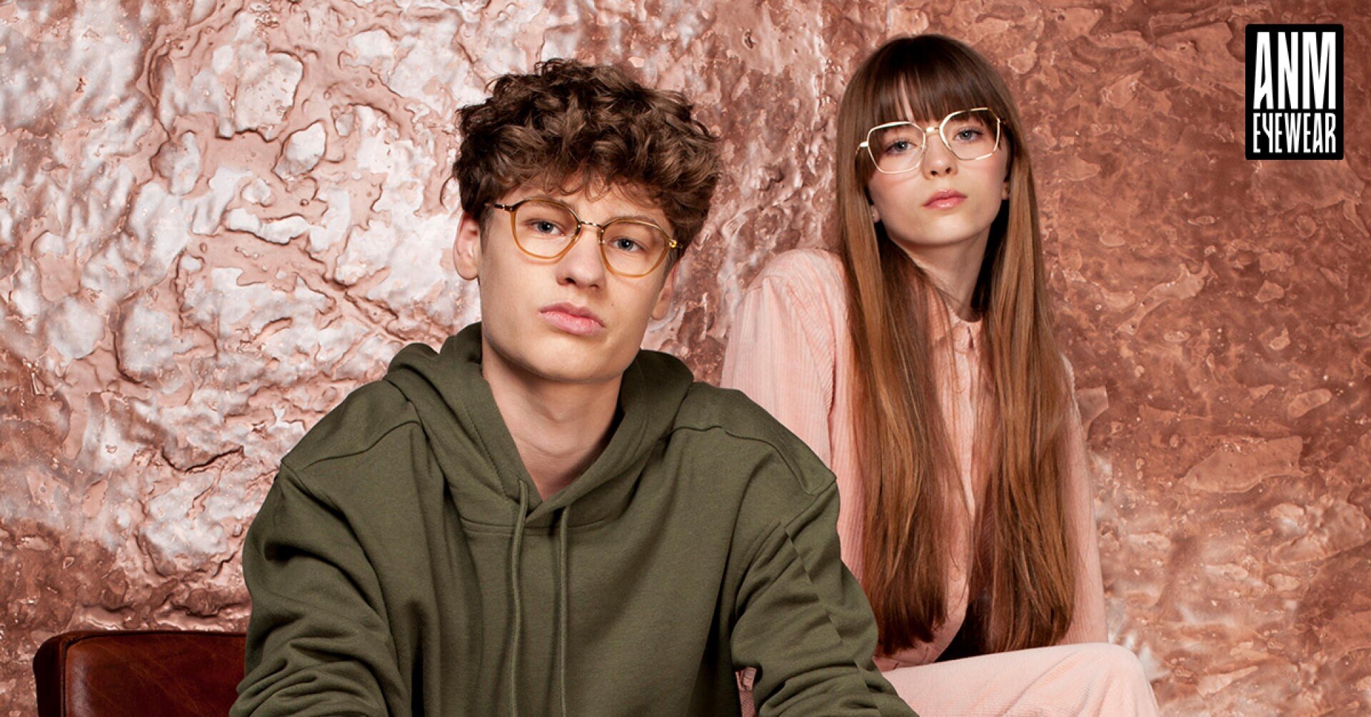 Ein junger Mann in einem grünen Kapuzenpullover und eine junge Frau in hellrosa Kleidung, beide tragen Brillen, sitzen vor einem kupferfarbenen, strukturierten Hintergrund mit dem ANM EYEWEAR-Logo in der oberen rechten Ecke.