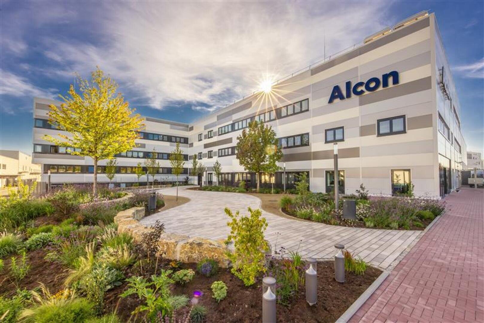 Modernes Bürogebäude mit "Alcon"-Schild, umgeben von Landschaftsgärten, Bäumen und einem gepflasterten Gehweg unter einem teilweise bewölkten Himmel mit Sonnenlicht, das über das Dach fällt.