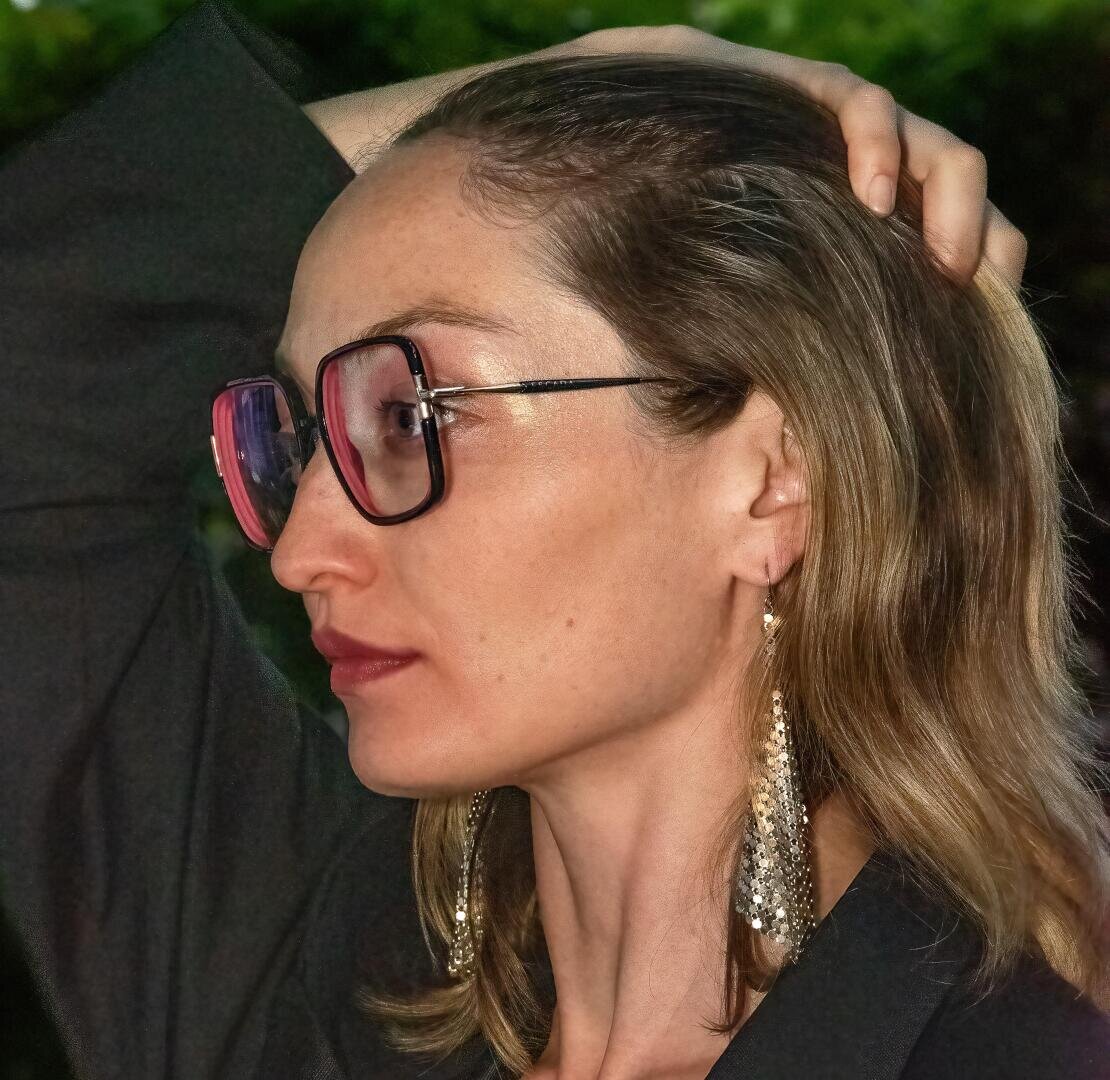 Eine Frau mit schulterlangem hellbraunem Haar, einer großen rosafarbenen Brille, langen goldenen Ohrringen und einem dunklen Oberteil blickt zur Seite, während sie ihr Haar mit einer Hand vor einem verschwommenen grünen Hintergrund zurückhält.