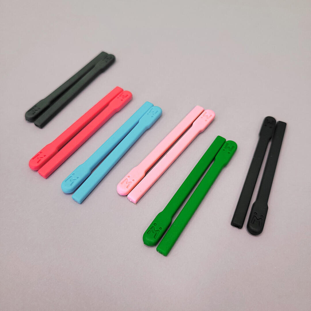 Acht bunte Kunststoffstäbchen, darunter schwarze, rote, blaue, rosa und grüne Paare, sind auf einer hellgrauen Oberfläche in zwei ungleichmäßigen Reihen angeordnet. Jedes Stäbchen hat ein kleines, geprägtes Gesicht in der Nähe der Spitze.