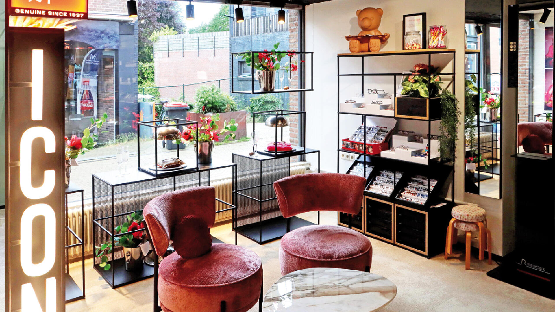 A modern, stylish shop interior with pink velvet chairs, glass display shelves holding plants and accessories, a marble table, and large windows letting in natural light. A sign reading ICON is illuminated on the left.