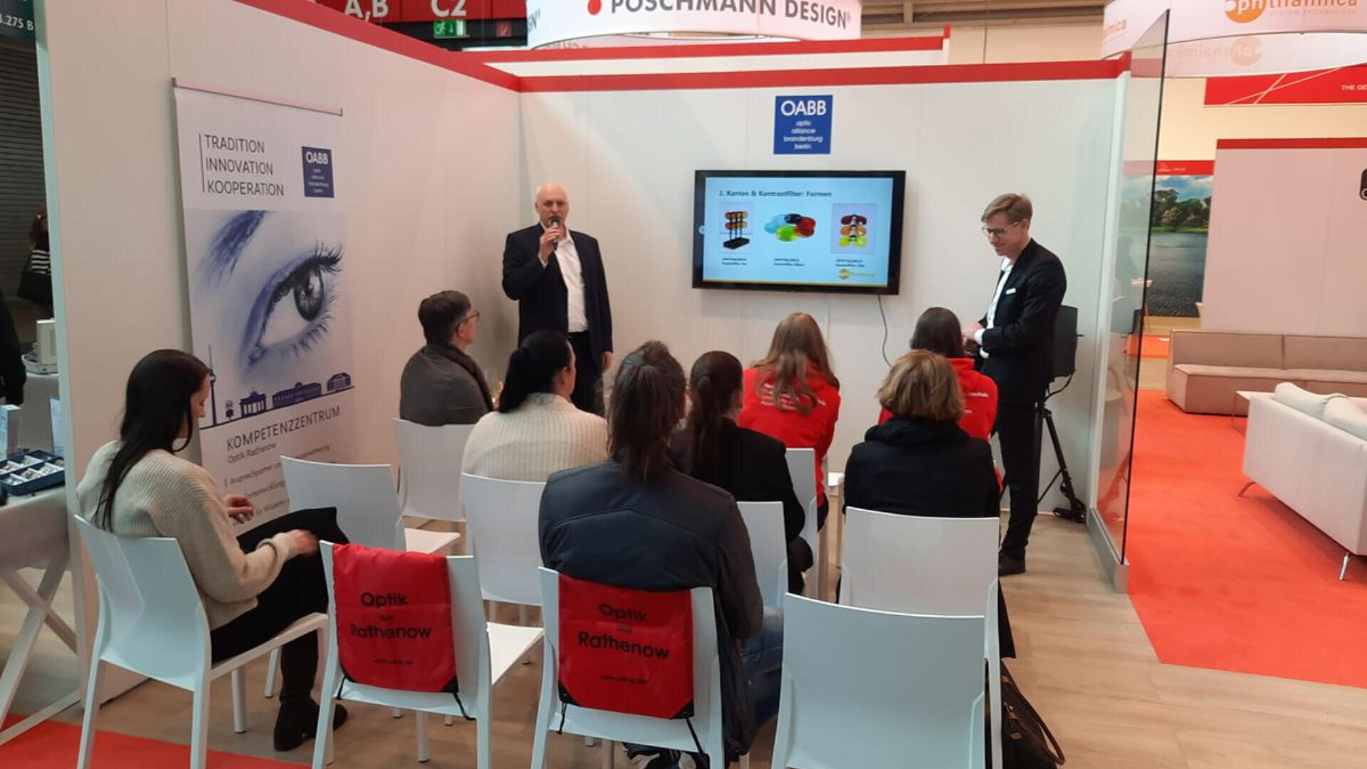 Ein Mann spricht in ein Mikrofon neben einem Bildschirm, auf dem eine Präsentation für ein sitzendes Publikum an einem Messestand läuft. Die meisten Zuschauer tragen rote Jacken mit der Aufschrift „Optik Rathenow“ auf dem Rücken.