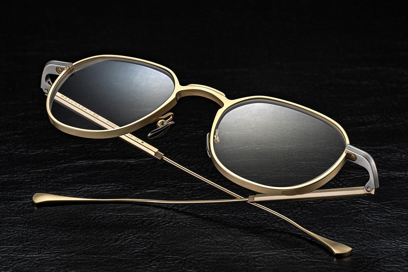 Gold-rimmed sunglasses with dark lenses are displayed on a black textured surface, with the arms crossed and the glasses facing upward.