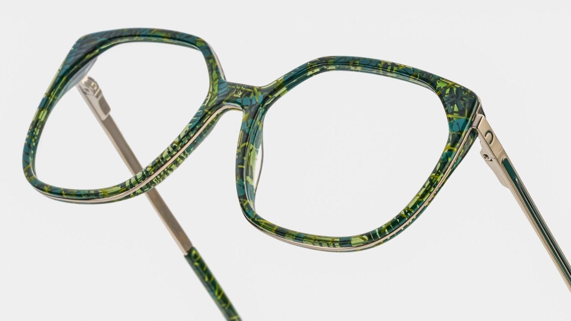 A close-up of stylish eyeglasses with large, geometric lenses and green, marbled frames. The temples are slim and metallic with green detailing at the hinges.