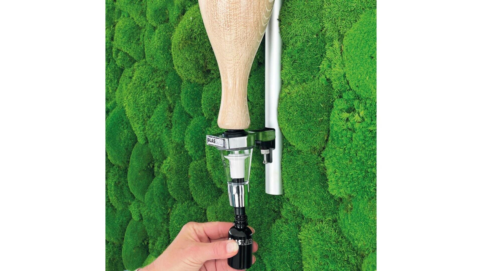 A hand attaches a prosthetic leg with a wooden top and metal components to a connector against a vibrant green moss-covered wall.