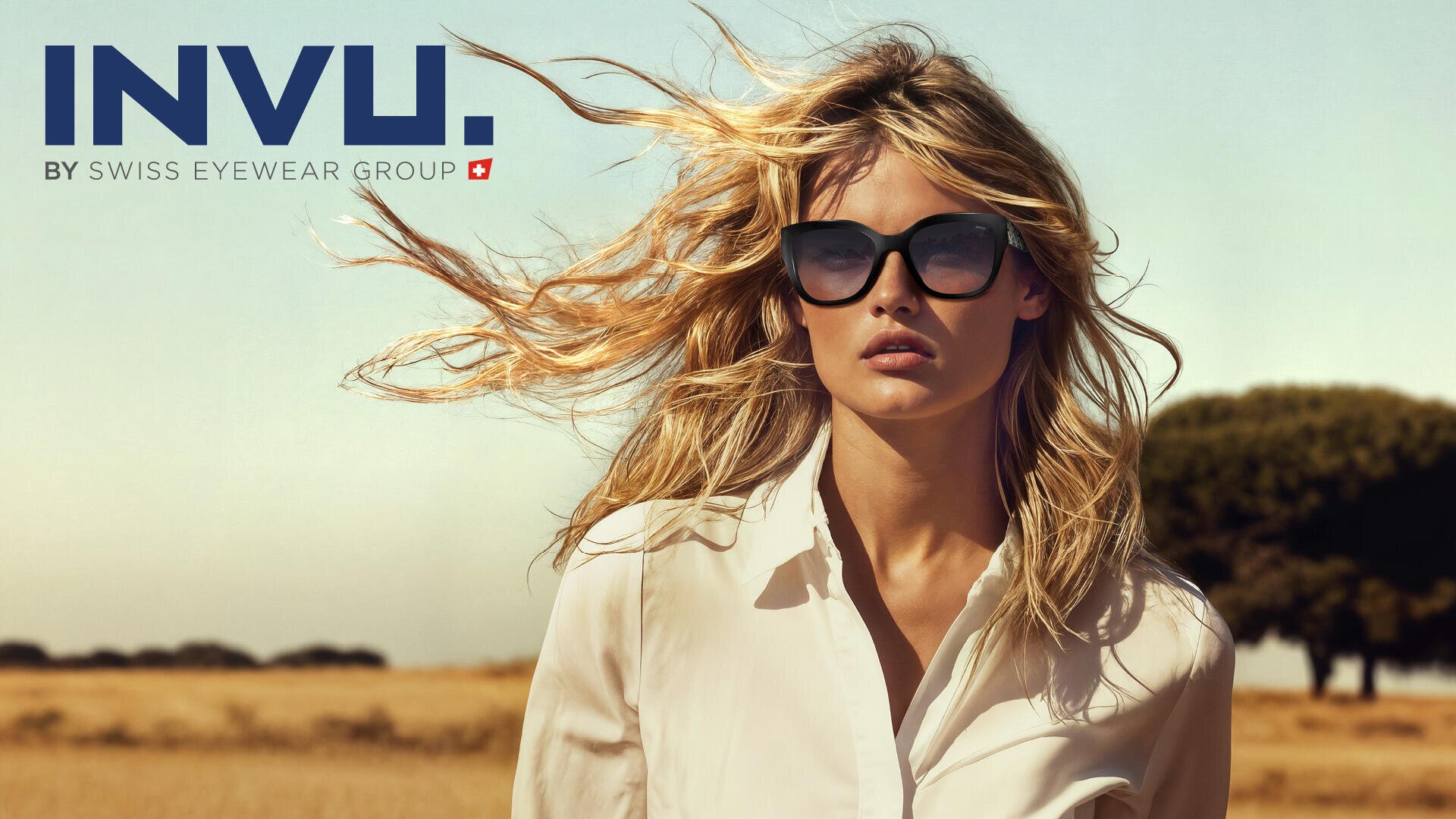 A woman with dark sunglasses and a white shirt is standing outside on a sunny meadow. The wind is blowing her blonde hair. The text reads INVU. by Swiss Eyewear Group in the top left corner.