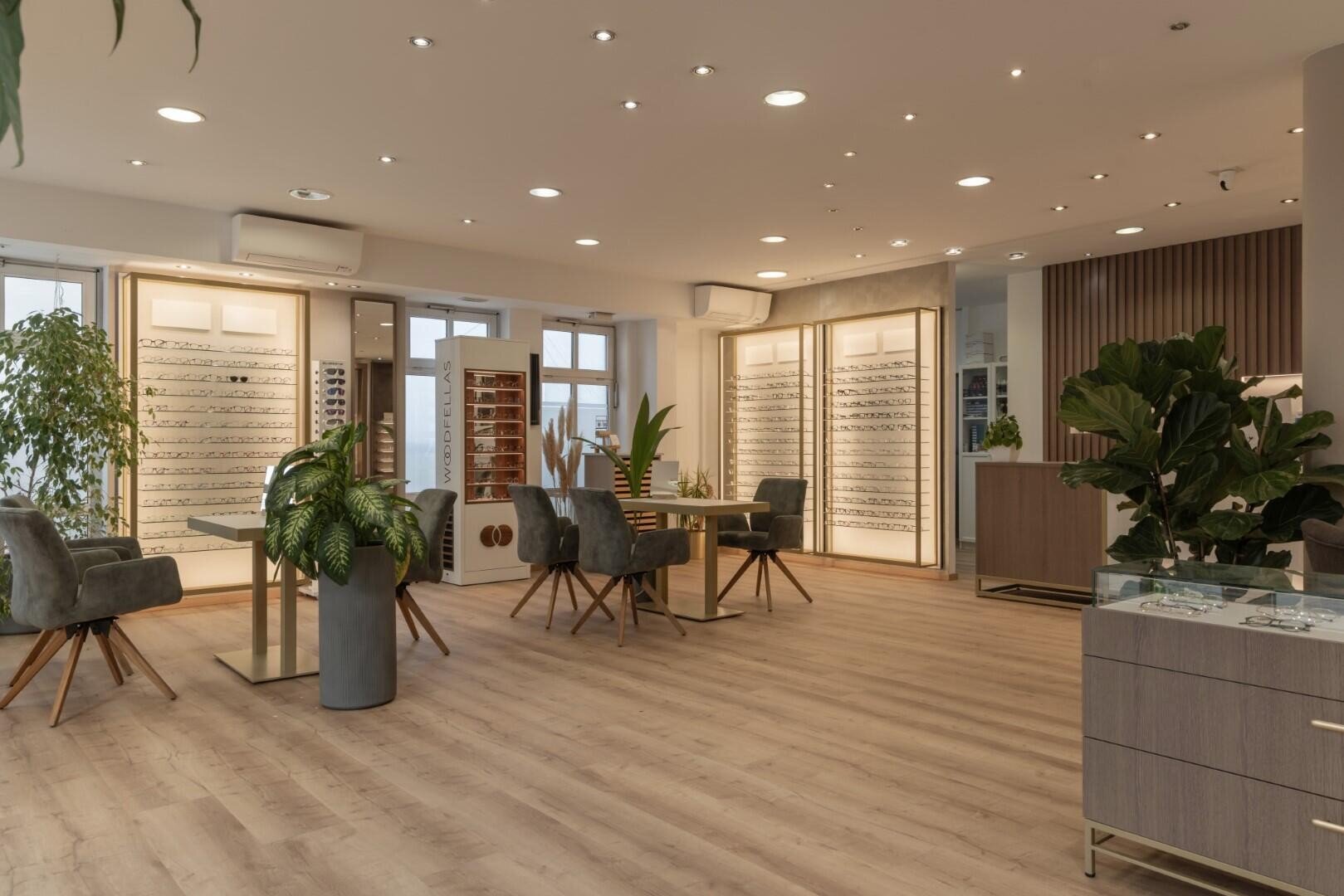 Ein modernes, geräumiges Optikergeschäft mit Holzboden, stilvollen Stühlen, Tischen, Brillenvitrinen und großen Zimmerpflanzen. Der Raum ist hell erleuchtet durch Deckenstrahler und Tageslicht durch große Fenster.