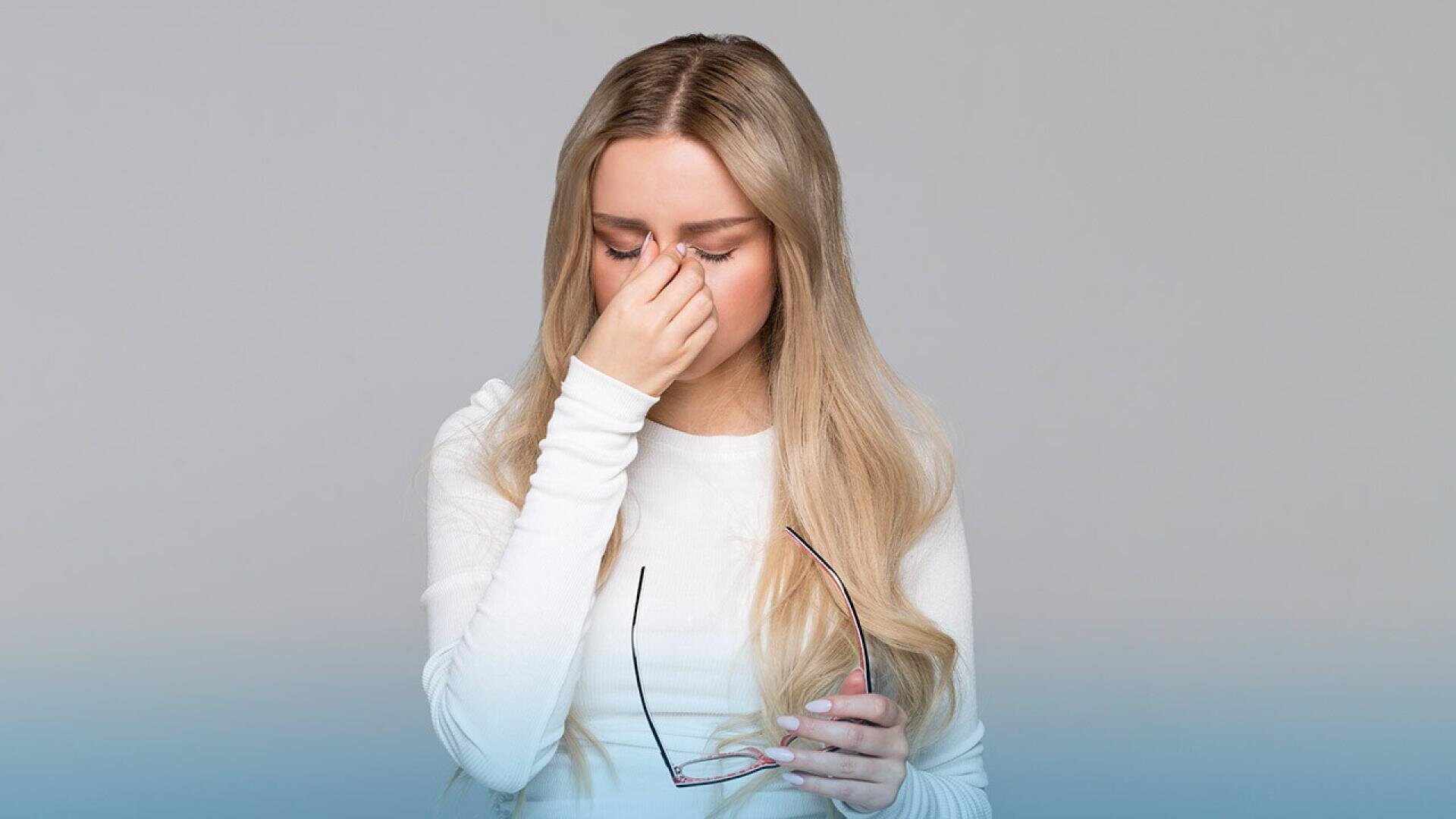A woman with long blonde hair in a white top holds eyeglasses in one hand and pinches the bridge of her nose with the other, appearing tired or stressed, against a plain light background.