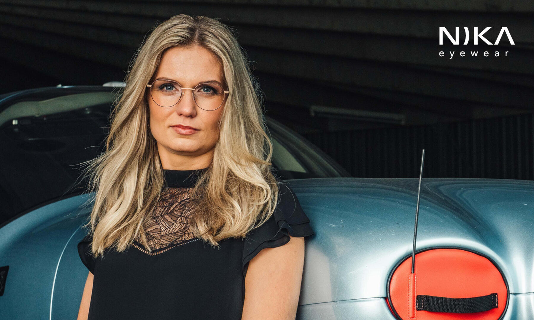 Eine Frau mit langen blonden Haaren und einer runden Brille steht neben einem hellblauen Auto. Sie trägt ein schwarzes Oberteil mit Spitzenmuster, und in der rechten oberen Ecke erscheint das NIKA-Brillenlogo.