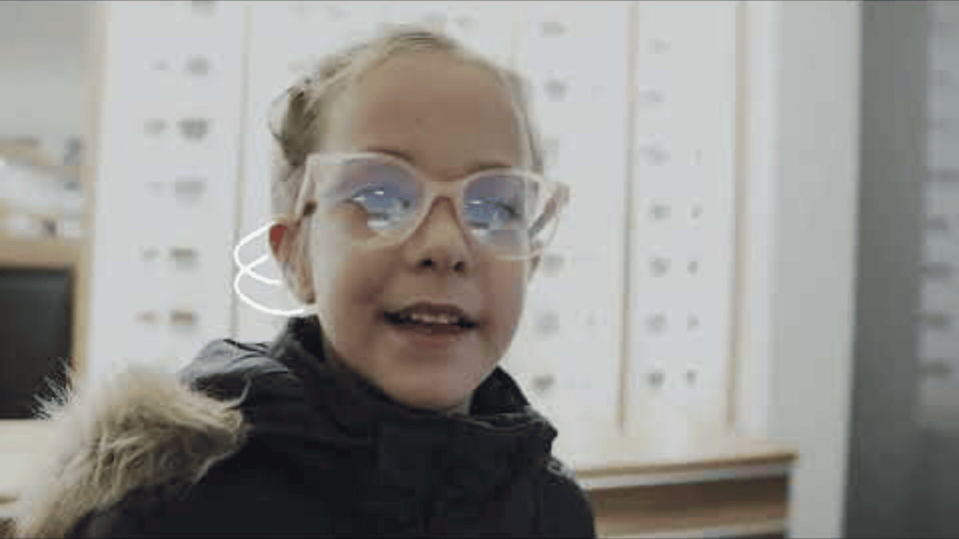 Ein junges Mädchen mit einer großen, durchsichtigen Brille und einem dunklen Mantel mit pelzgefütterter Kapuze lächelt im Innenbereich, vor einem unscharfen Hintergrund sind Reihen von Brillengestellen zu sehen.