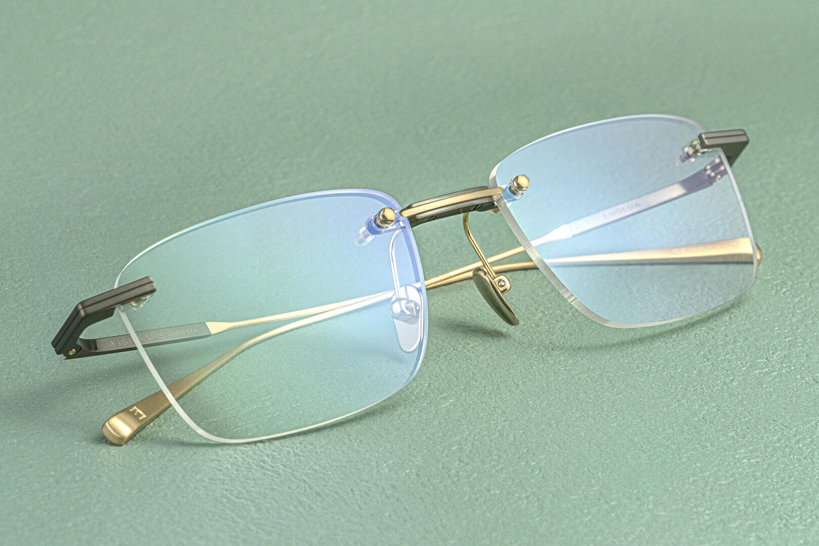 A pair of rimless eyeglasses with gold temples and blue-tinted lenses rests on a textured green surface.