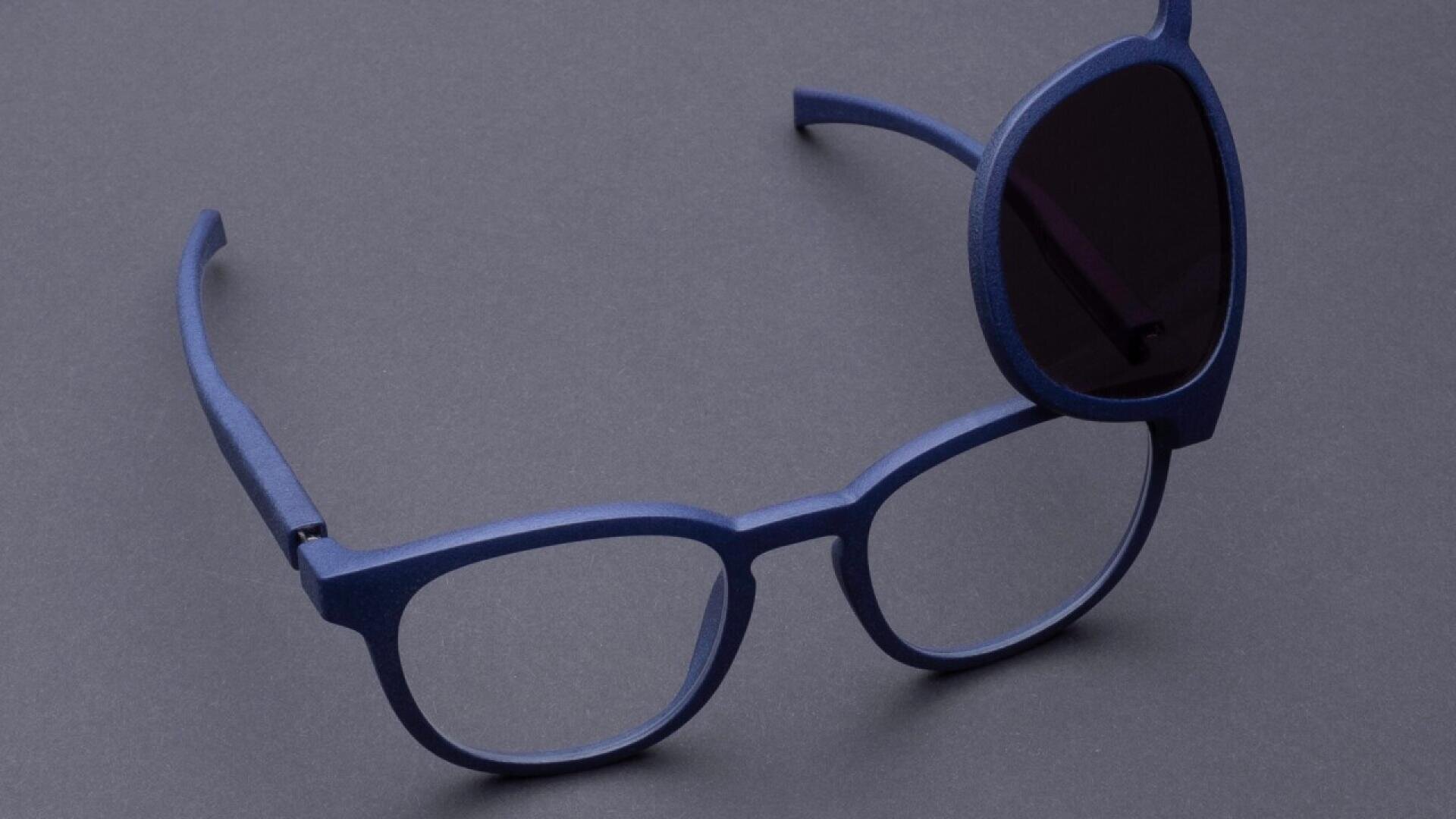 Blue eyeglasses with a detachable round magnetic sunglasses lens partly attached over the right lens, placed on a gray surface.