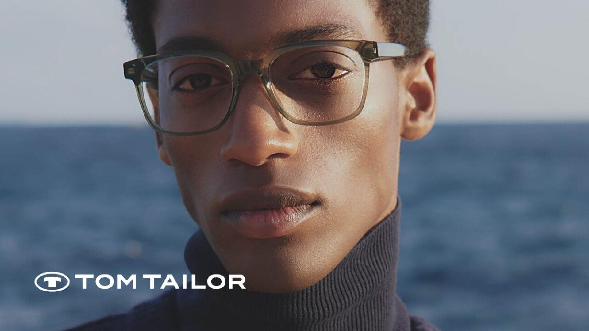 Close-up of a person wearing square glasses and a dark turtleneck, standing in front of a blurred blue ocean background. The Tom Tailor logo appears in the lower left corner.