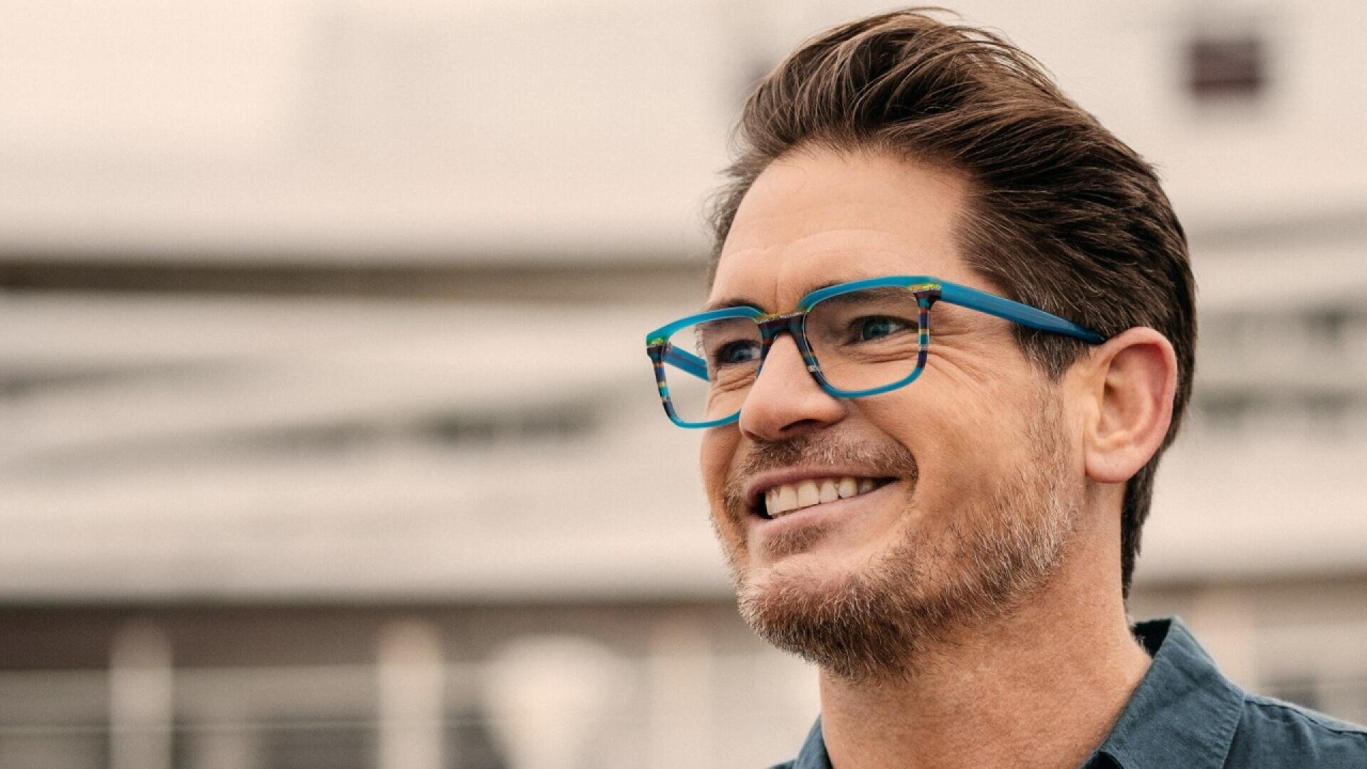 A man with short brown hair and a trimmed beard smiles while wearing blue glasses and a dark shirt, standing outdoors with a blurred background.