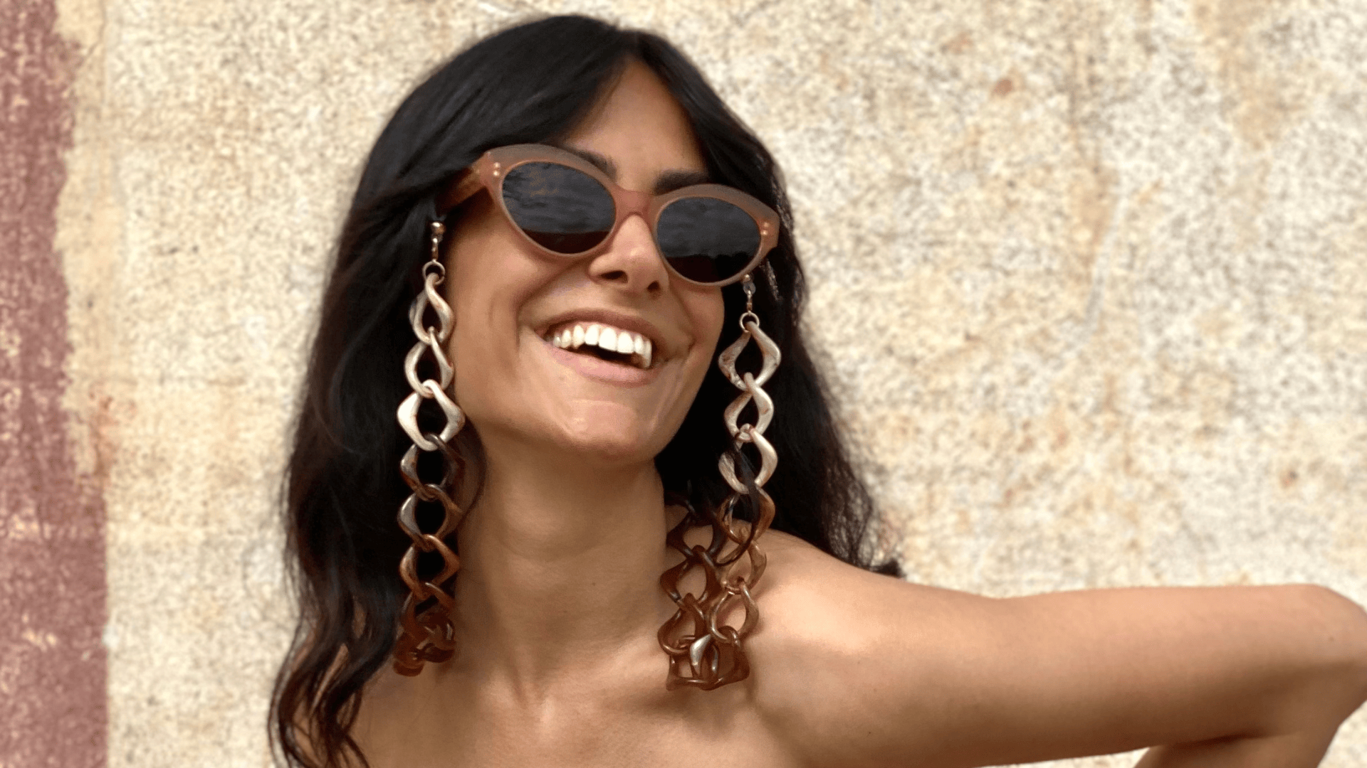 A smiling woman with long dark hair wears large sunglasses and oversized, chain-link earrings. She stands in front of a textured stone wall and looks off to the side, radiating joy.