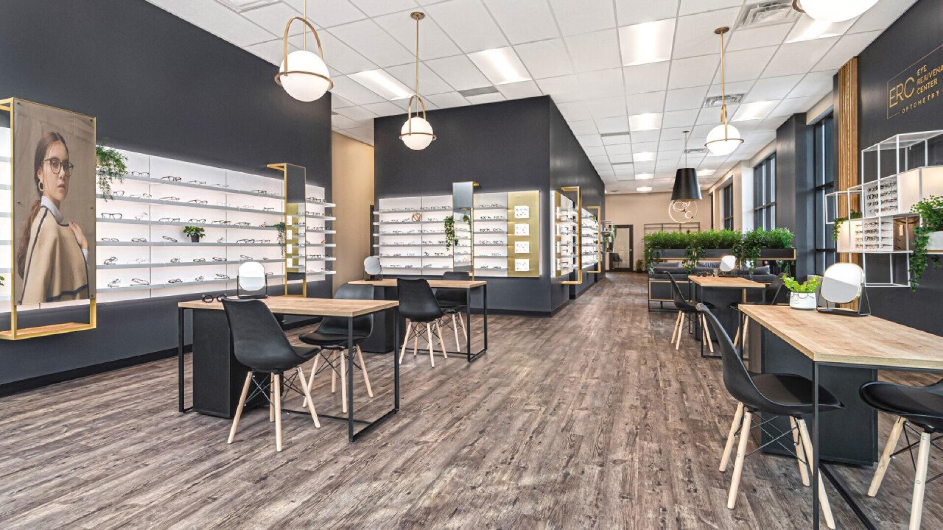 Modern optometry store with tables and chairs for consultations, shelves displaying eyeglass frames, potted plants, large windows, wood flooring, and bright lighting. A portrait of a woman in glasses is on the left wall.
