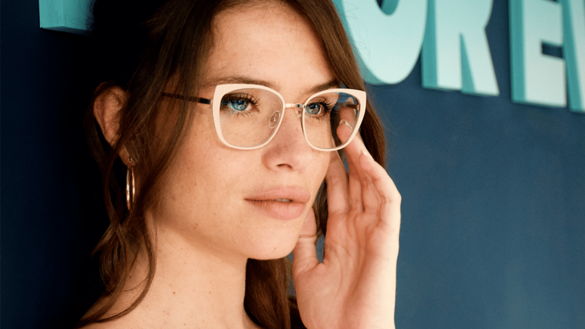 A woman with light brown hair and blue eyes wearing large clear-framed glasses and gold hoop earrings looks thoughtfully ahead while touching her glasses. Blue letters are visible in the background.