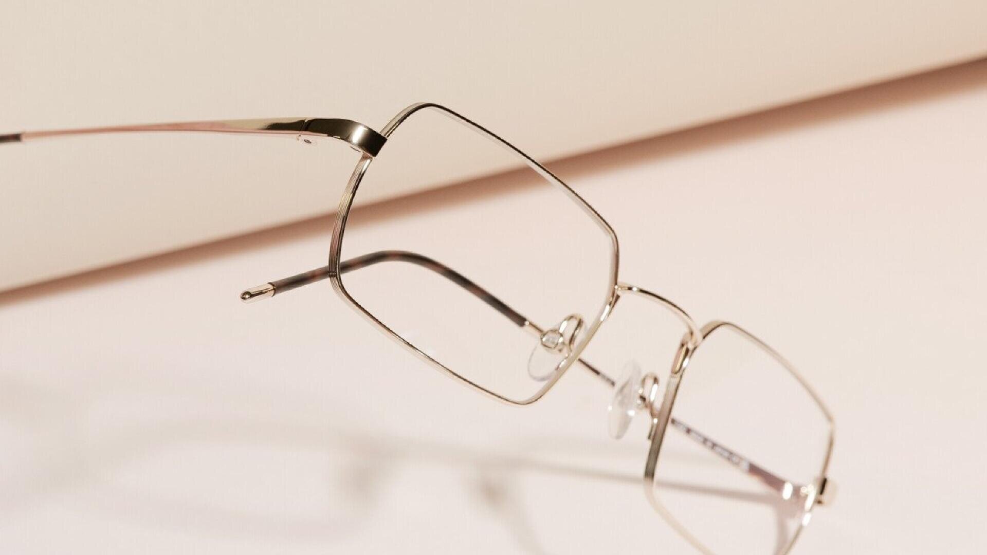 A pair of thin, silver metal eyeglasses with geometric frames resting on a light, neutral surface with a soft, blurred background.