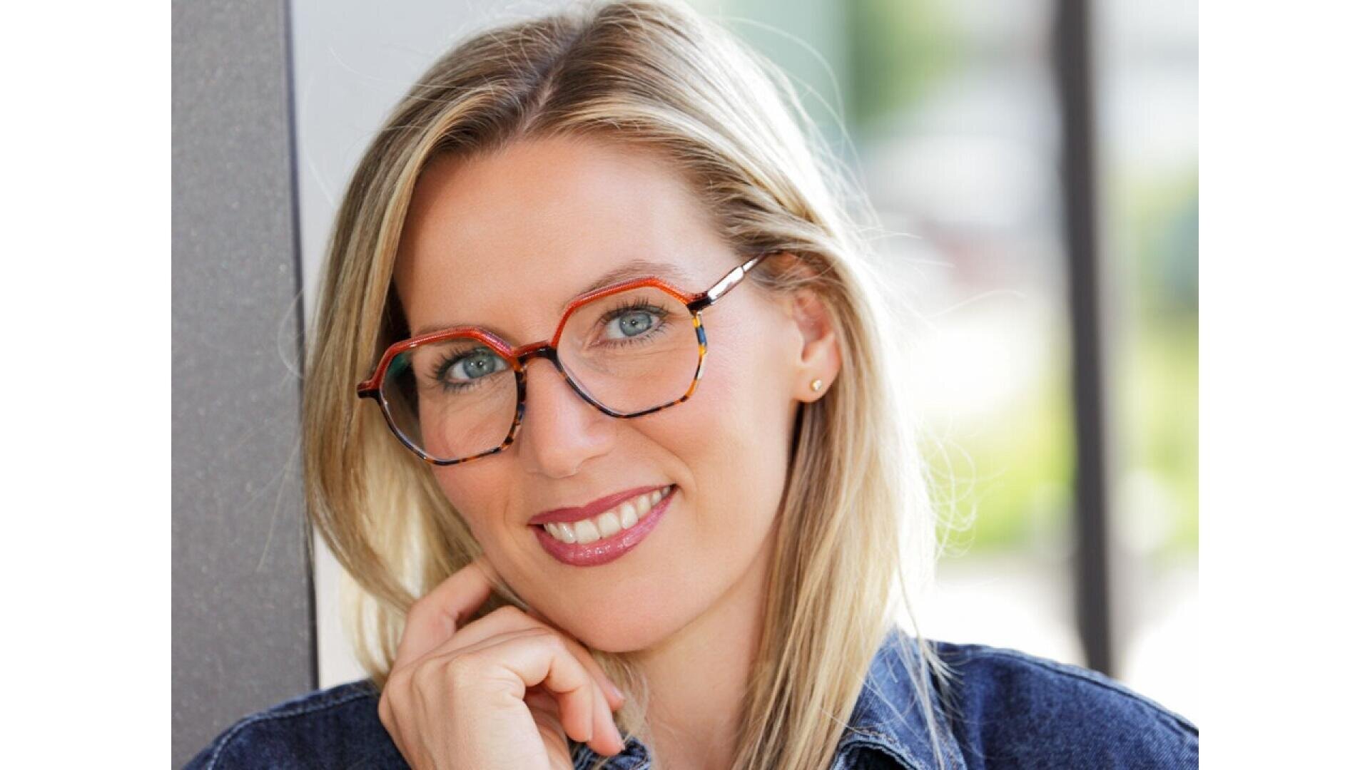 A smiling woman with long blonde hair and red-framed glasses rests her chin on her hand. She is wearing a denim jacket and stands in front of a blurred background.