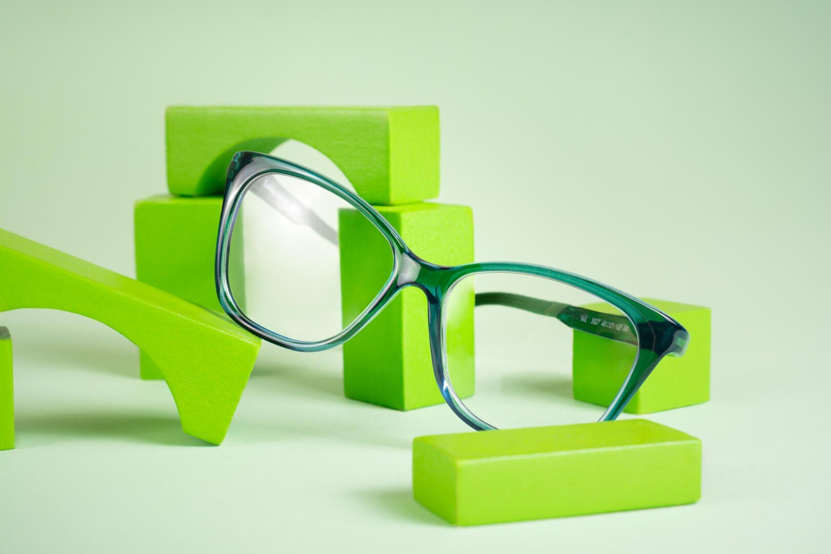 Zwischen verschiedenen hellgrünen Holzklötzen auf hellgrünem Hintergrund ist eine Brille mit grünem Rand ausgestellt.