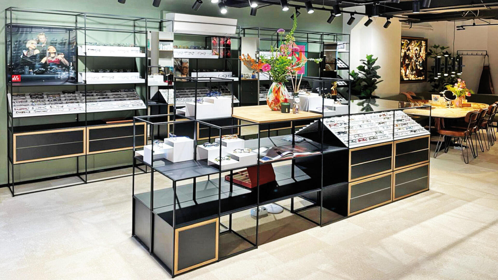 Modern eyewear store interior with black and gold display shelves holding various eyeglass frames, a central wooden counter with colorful flower arrangements, and seating area in the background. The space is brightly lit and organized.