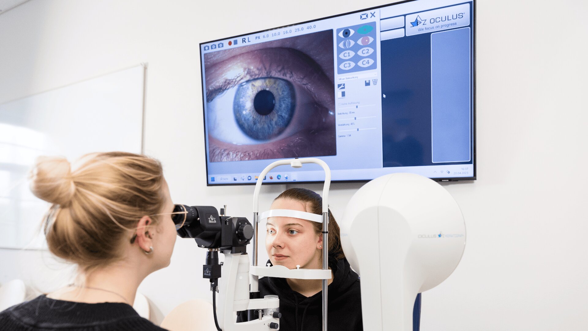 Eine Frau wird mit einem Spezialgerät einer Augenuntersuchung unterzogen, während ein Optiker das Gerät bedient. Auf einem großen Monitor wird eine Nahaufnahme des Auges der Patientin im Detail angezeigt.