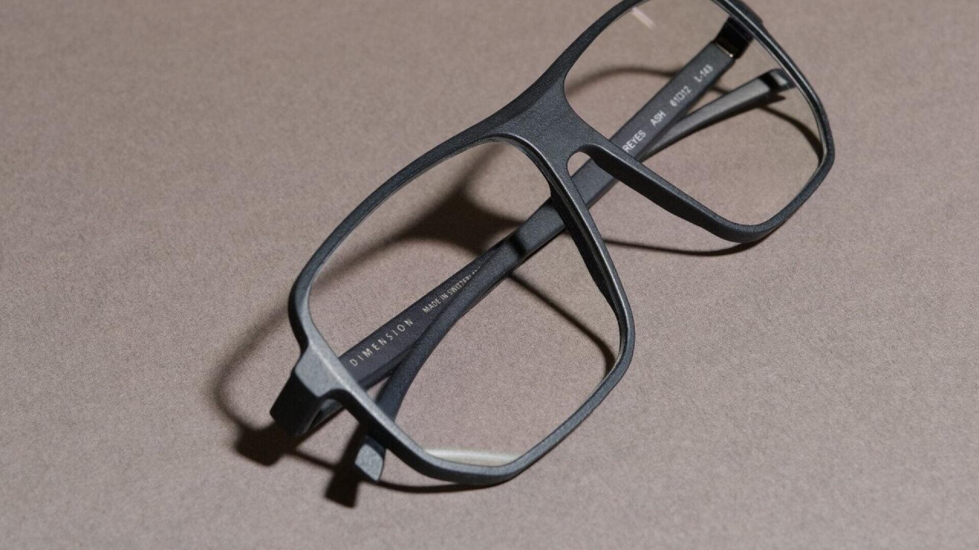 A pair of black rectangular eyeglasses with thick frames lies on a flat, light brown surface, casting a slight shadow.