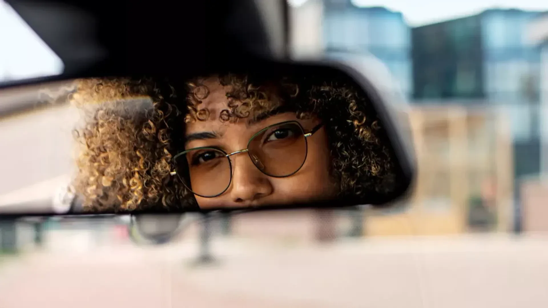 A close-up of a persons face with curly hair and glasses, reflected in a cars rearview mirror. The background outside the car window is blurred.
