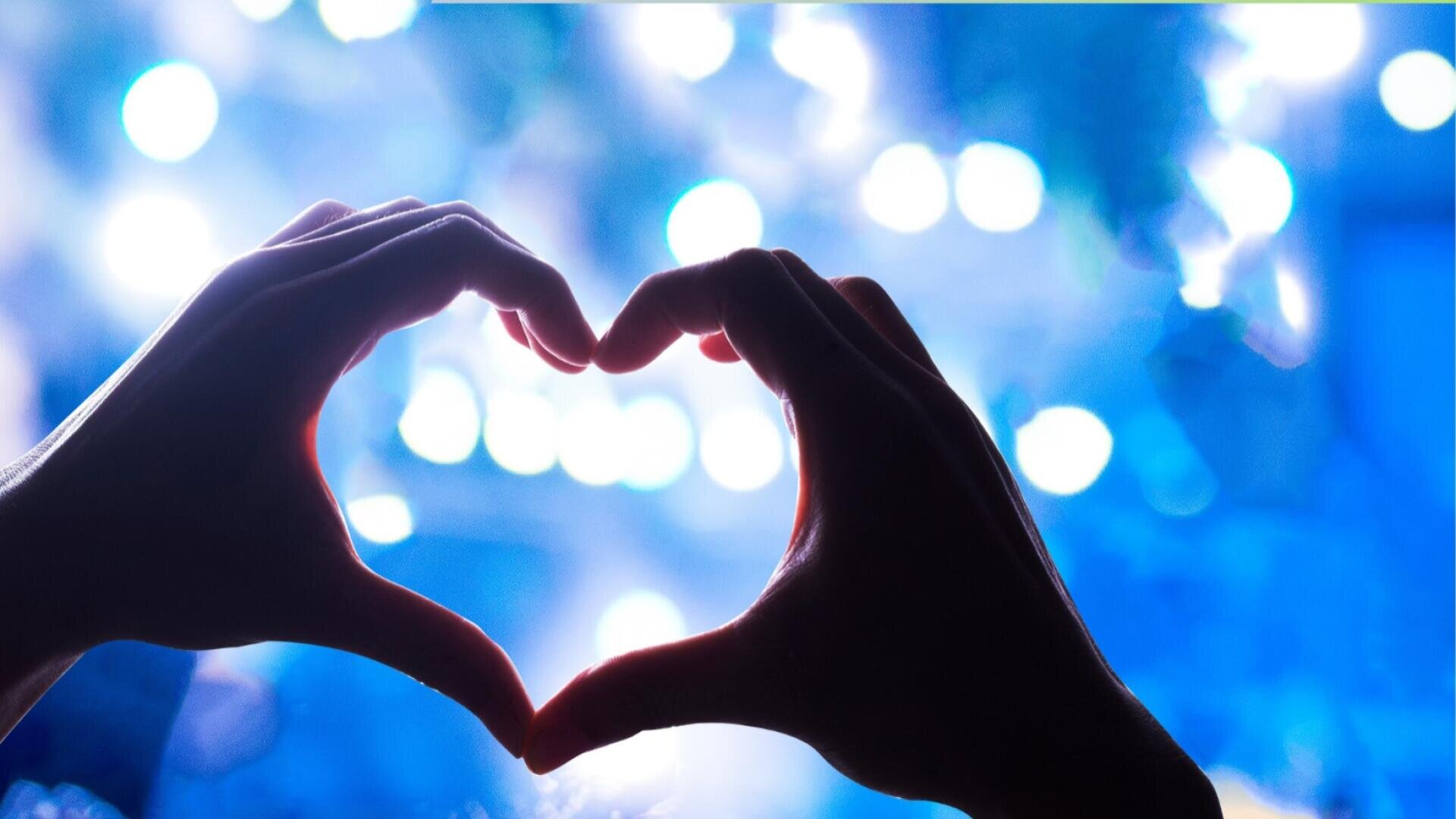 Zwei Hände bilden die Form eines Herzens vor einem Hintergrund aus blauen Lichtern, die eine sanfte, leuchtende und romantische Atmosphäre schaffen.
