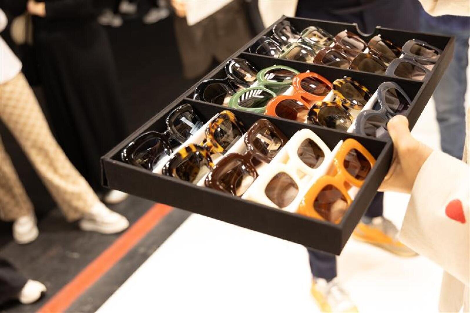 Eine Person hält ein Tablett mit verschiedenen bunten Sonnenbrillen mit unterschiedlichen Fassungen und Farben. Im unscharfen Hintergrund stehen mehrere Personen, einige tragen gemusterte Hosen und Turnschuhe.