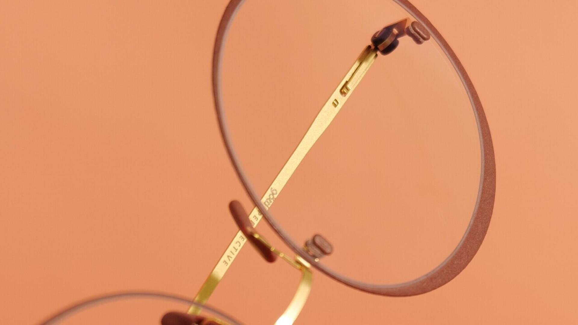 Close-up of round, gold-framed eyeglasses with brown rims, positioned against a peach-colored background. The nose pads and part of the temple arm are visible.