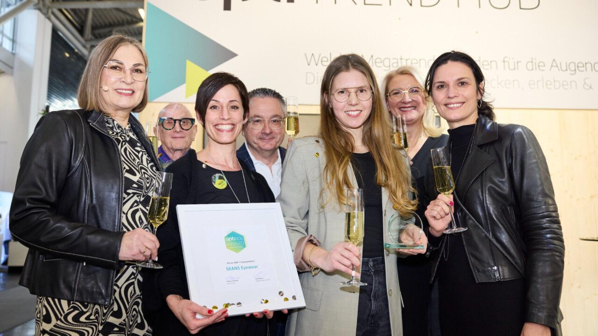 Six people stand together indoors, smiling and holding champagne glasses. One woman in front holds a framed award. A TREND HUB sign and modern decor are visible in the background.