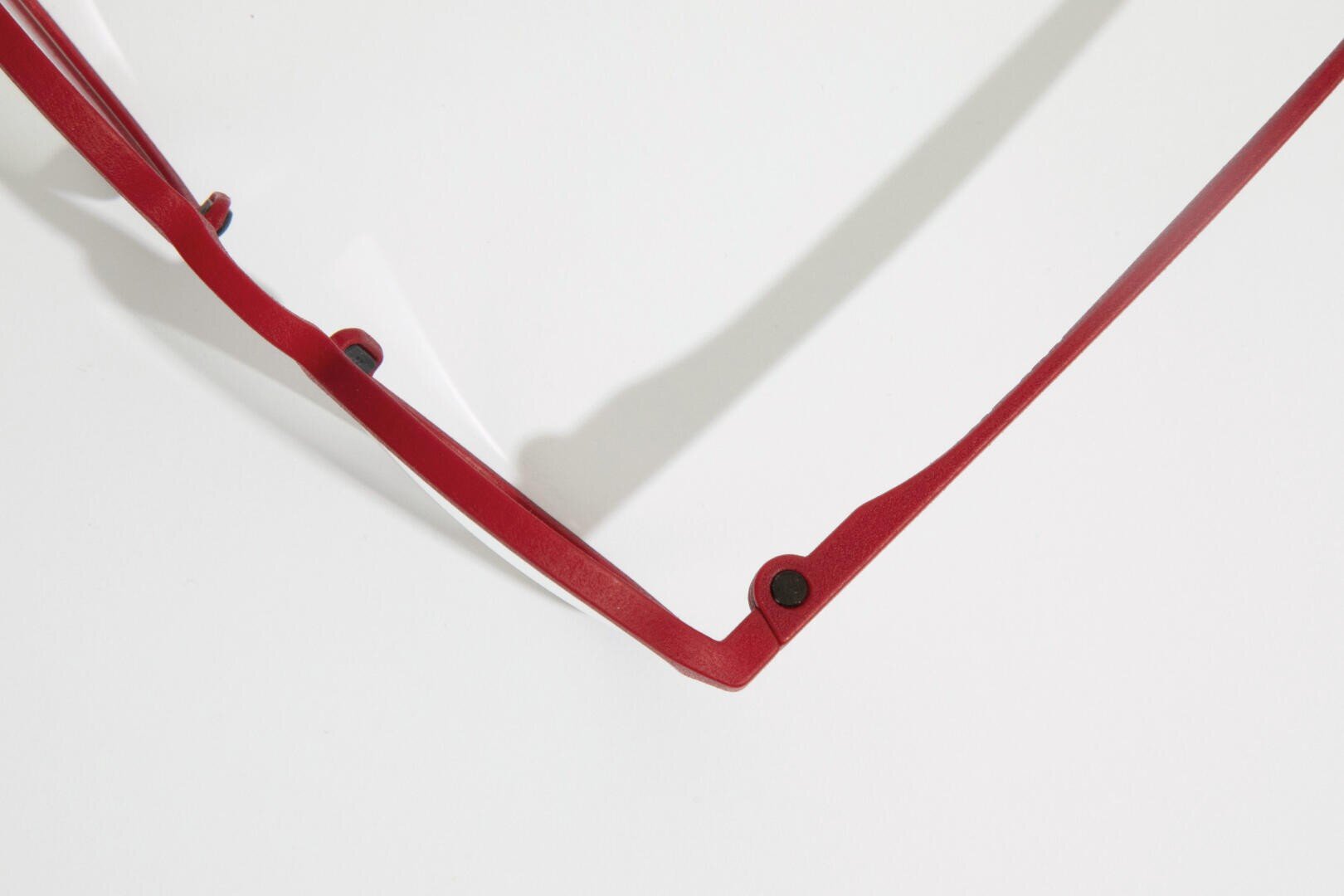 Nahaufnahme eines roten Brillengestells mit Schwerpunkt auf dem Scharnier und dem Bügel vor einem weißen Hintergrund. Das Bild ist minimalistisch, zeigt nur einen Teil des Rahmens und wirft einen weichen Schatten.