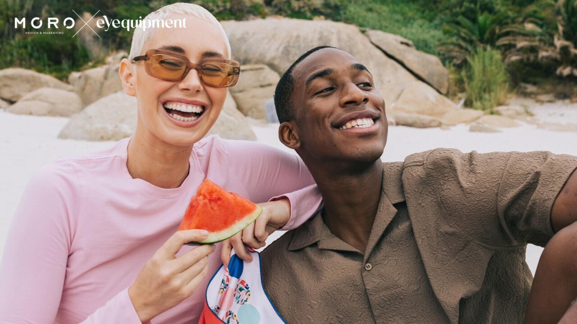 Two friends sit on a beach, smiling and enjoying a sunny day. One person holds a slice of watermelon while wearing sunglasses and a pink top; the other wears a brown shirt. Large rocks and greenery are visible in the background.