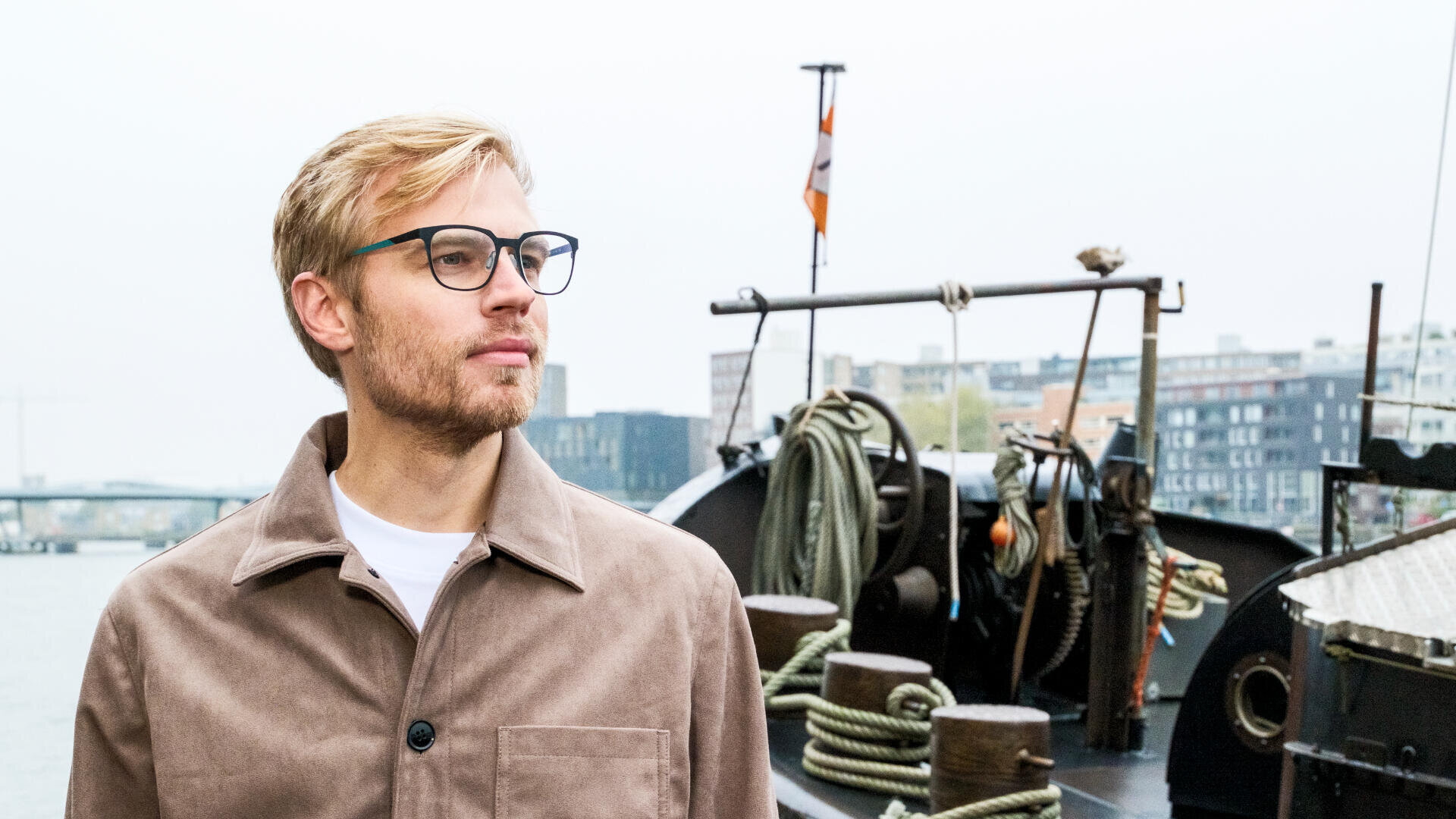 Ein Mann mit blondem Haar und Brille, der eine beige Jacke trägt, steht vor einem angedockten Boot mit Seilen und Ausrüstung, im Hintergrund sind an einem bewölkten Tag Gebäude zu sehen.