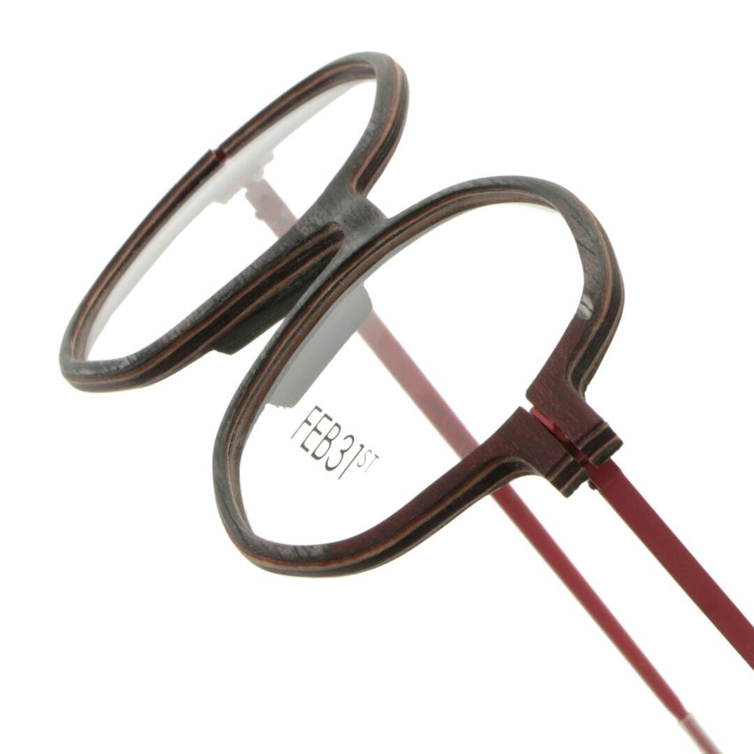Ovale Brille mit braunem Rahmen mit Holzstruktur und roten Bügeln. Auf der Innenseite des rechten Glases ist der Text FEB31st aufgedruckt.