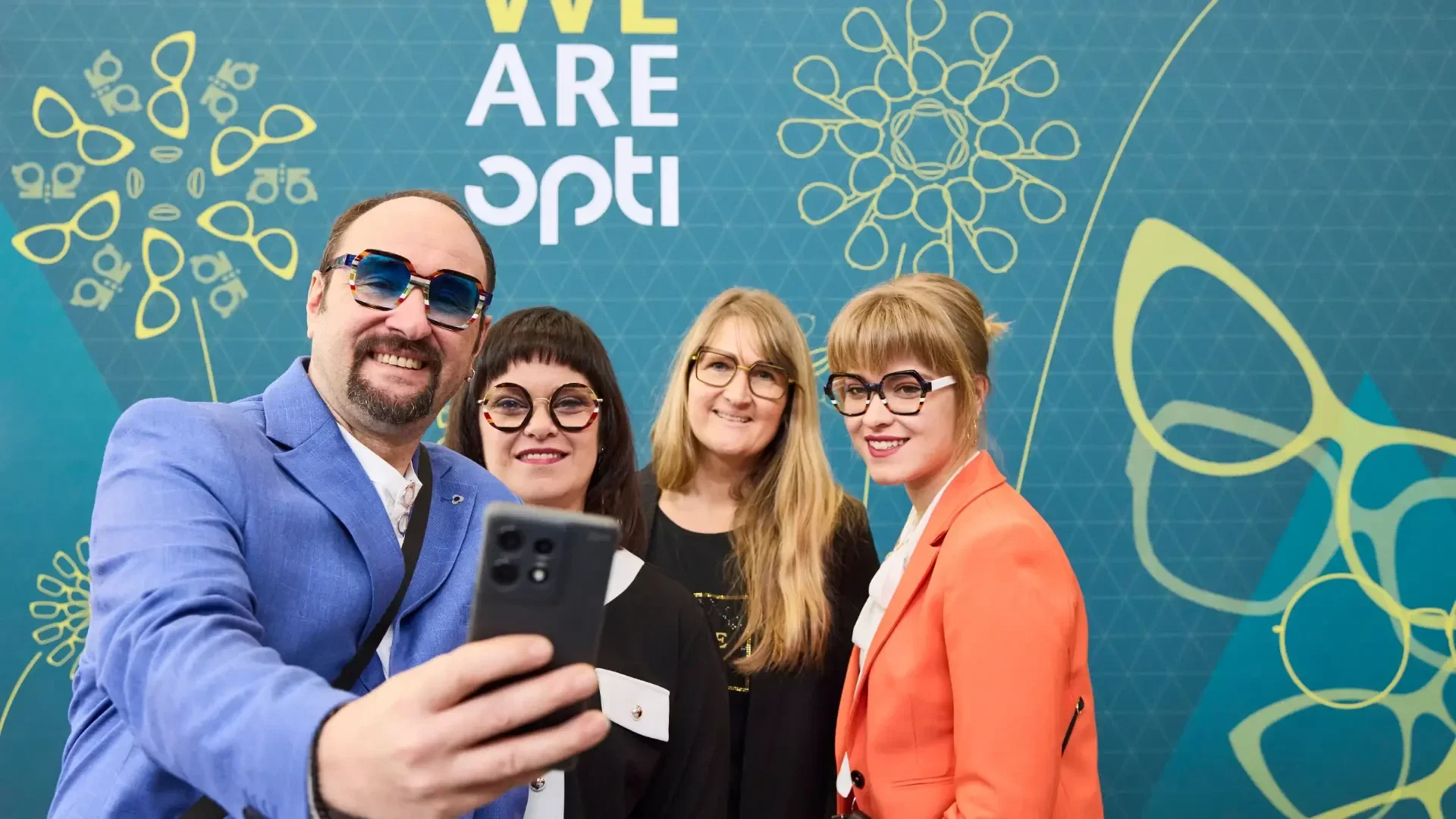 Four people wearing stylish glasses pose for a selfie in front of a blue backdrop with yellow designs and the text WE ARE opti. They are smiling and appear to be at an event or exhibition.