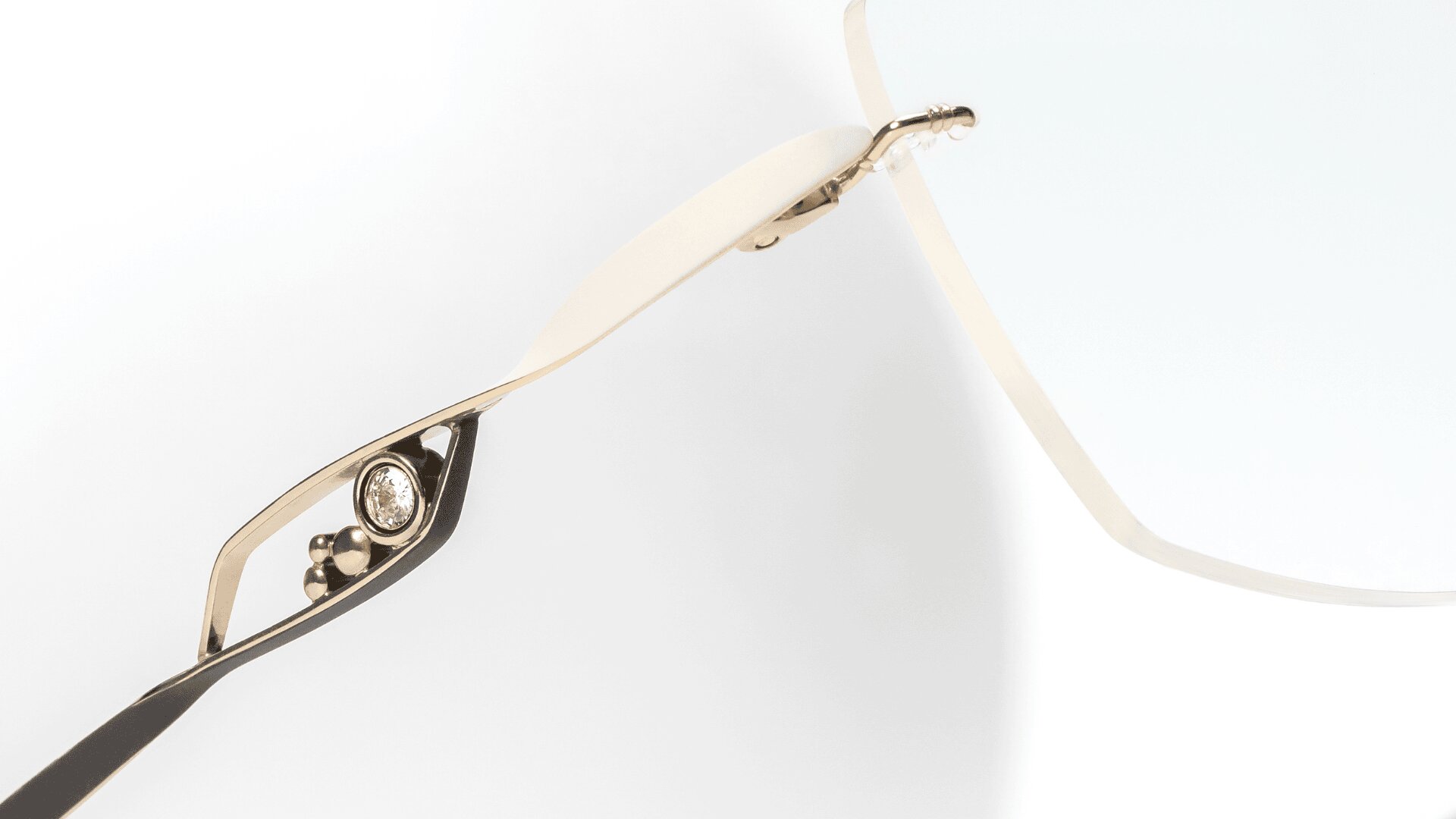 Nahaufnahme der Seite einer Brille mit goldenem Rahmen, mit einem dekorativen Ausschnitt und einem kleinen runden Edelstein in der Nähe des Scharniers. Der Hintergrund ist hell und minimalistisch.