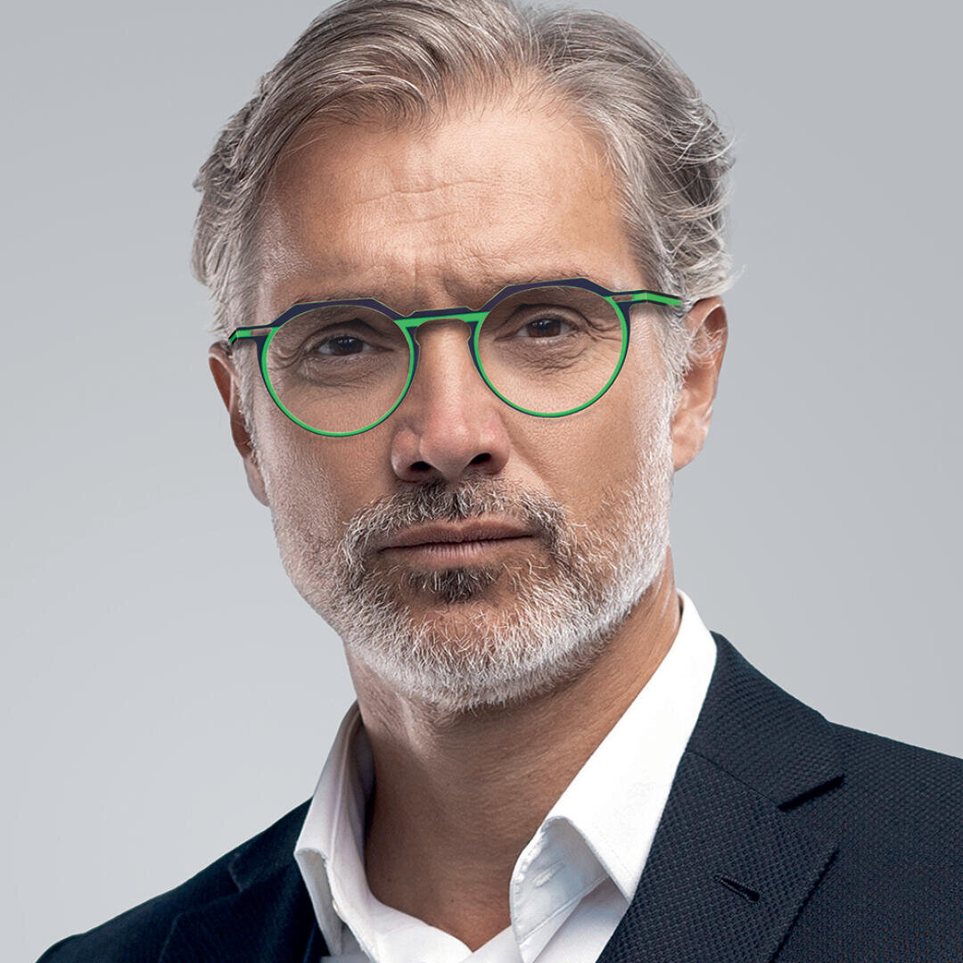 Ein Mann mittleren Alters mit grauem Haar, kurzem Bart und Schnurrbart trägt eine grün umrandete Brille und ein dunkles Jackett über einem weißen Hemd und steht vor einem schlichten grauen Hintergrund.