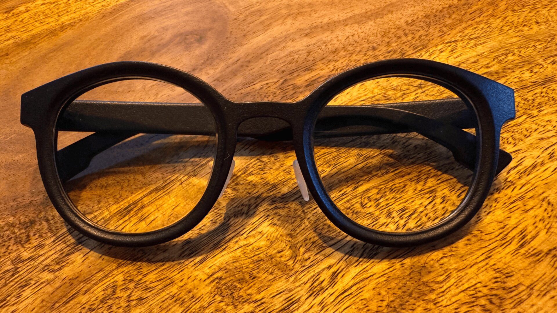 A pair of black, round-framed eyeglasses with thick rims rests on a wooden surface with a warm, textured grain.