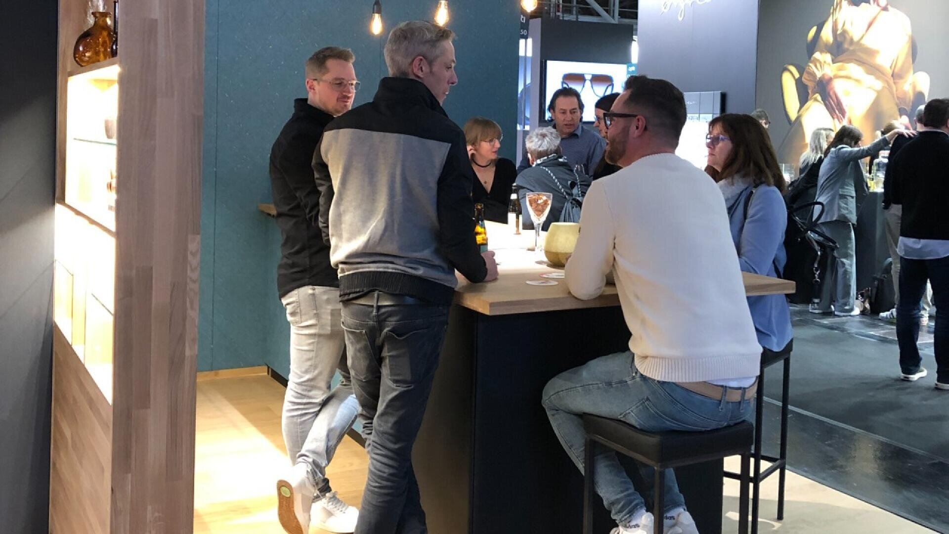A group of people sit and stand around a high table in a modern indoor setting, talking and drinking. The atmosphere is casual, with contemporary decor and hanging lights in the background.