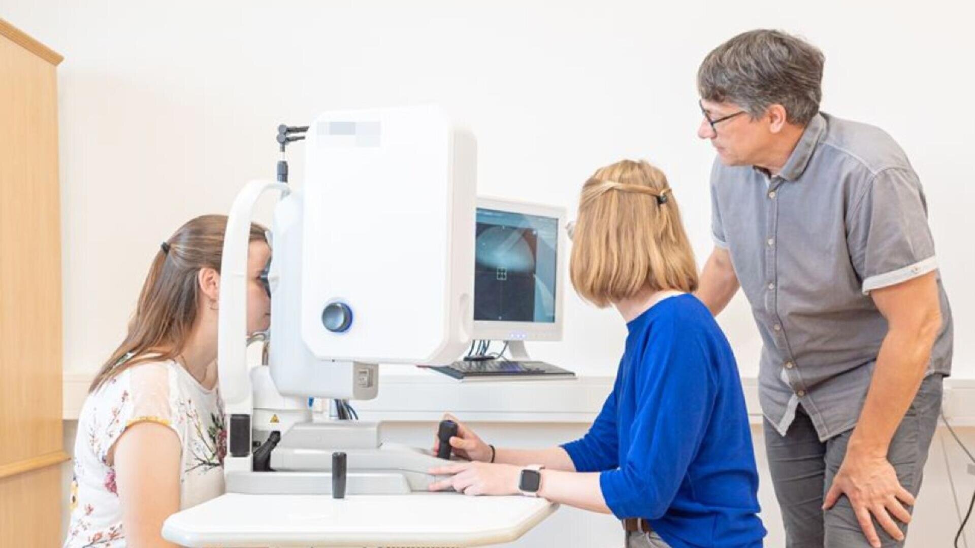 A woman sits at an eye examination machine while another woman operates it, and a man stands by observing; a computer screen displays the eye test results in a clinical setting.