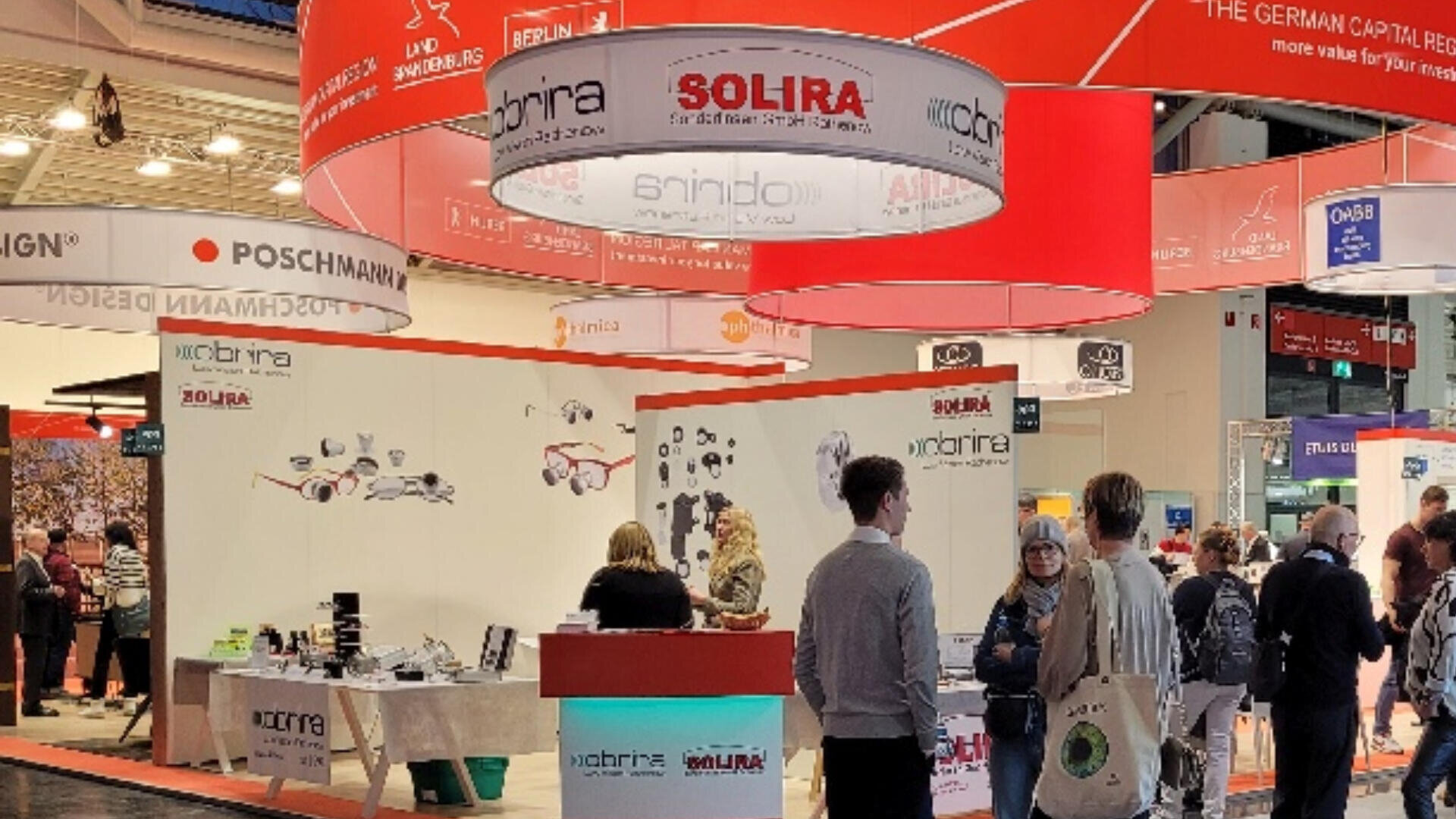 People visit a busy exhibition booth for SOLIRA at a trade show, with red signage overhead, white walls, product displays, and informational materials; several attendees interact with staff at the booth.